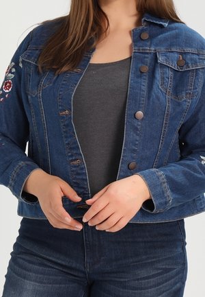 Kvinde knapper en blå denimjakke med blomsterbroderi på ærmerne, iført mørkegrå top og mørkeblå jeans.