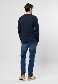 Camisetta navy a maniche lunghe con scollatura rotonda, abbinata a un paio di jeans blu. La persona indossa sneakers beige, in piedi contro uno sfondo chiaro uniforme.