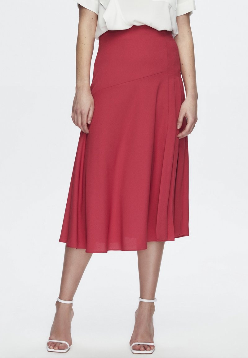 Falda roja hasta las rodillas hecha de un tejido ligero con diseño plisado. Presenta una cintura ajustada y una textura suave, combinada con sandalias blancas de tiras.