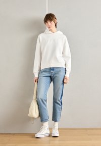 Felpa bianca con testo in rilievo, jeans azzurri chiari con bande laterali e scarpe sportive bianche alte. Una borsa crema è tenuta in una mano.