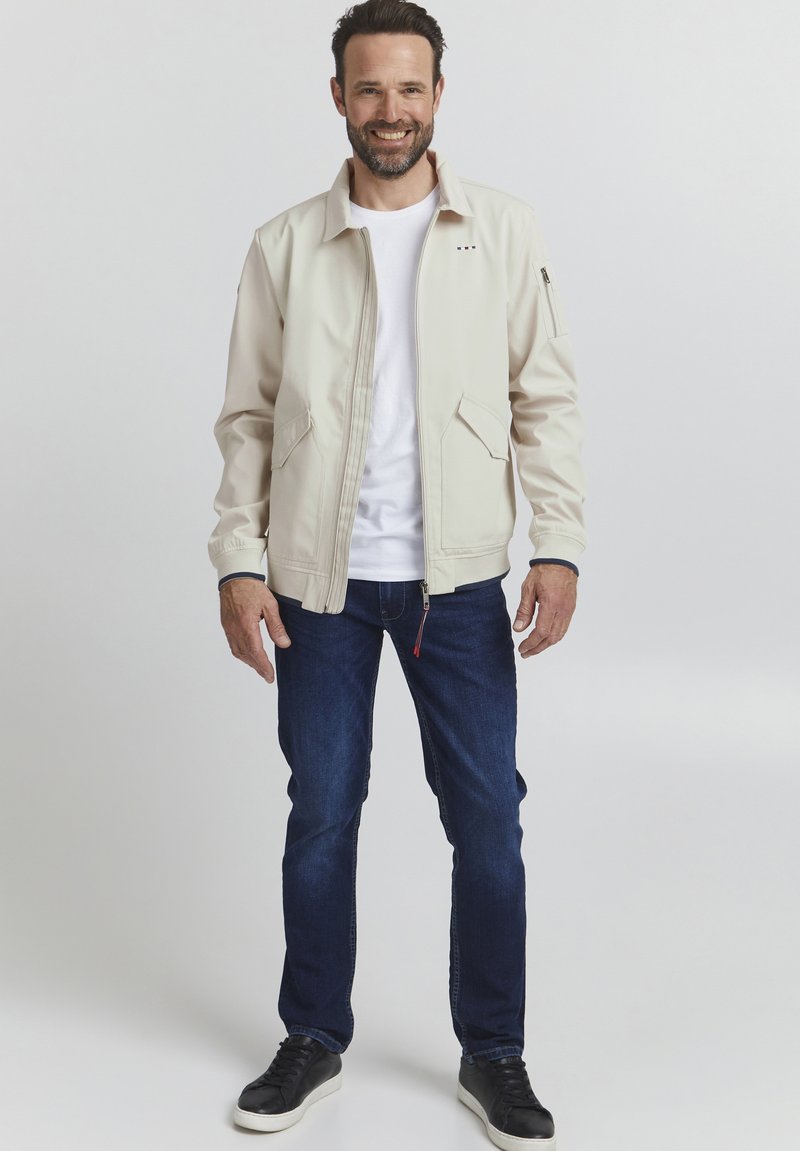 Light beige bomber jacket with a zip front, two side pockets, ribbed cuffs, and a white T-shirt underneath; worn with dark blue jeans and black sneakers.
