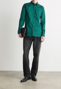 Chemise verte à manches longues, coupe ajustée, avec col. Associée à un jean foncé et des chaussures noires. Tenant une pochette noire.