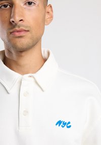 Polo shirt bianco in cotone con colletto a pieghettatura, caratterizzato da una texture leggera e da un logo "NYC" ricamato in blu sul petto.