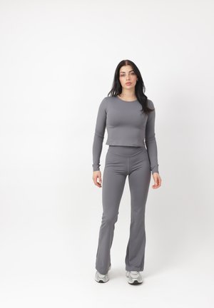 Kvinde stående iført tætsiddende grå langærmet top, matchende højttaljede bukser og hvide sneakers mod hvid baggrund.