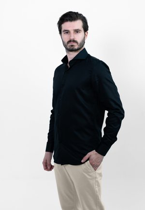 Zwarte lange mouwen button-up shirt met een gladde textuur en puntige kraag, gecombineerd met lichtbeige broek. Geen opmerkelijke patronen of accenten.