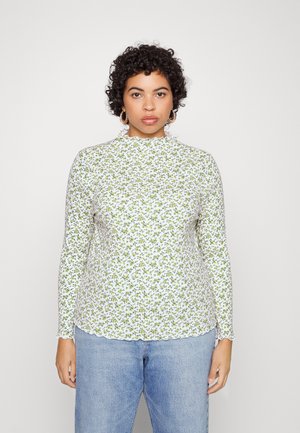 Haut à manches longues blanc avec un motif floral vert, présentant un col volanté et un ourlet en vagues, associé à un jean bleu clair.