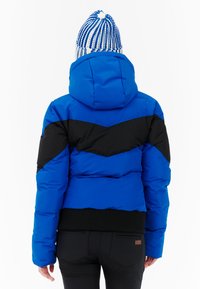 Modrá puffová bunda s čiernym chevronovým vzorom, s kapucňou a elastickými čiernymi manžetami. Pletená modro-biela čiapka na hlave modela.