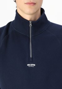 Navy blå hettegenser med glidelås og høy krage, laget av myk stoff. Har sølvfarget glidelås og hvit "Hollister" logo på brystet.