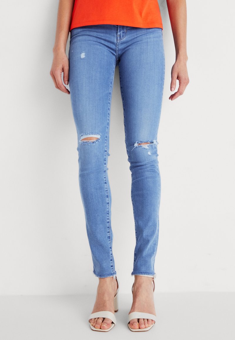 Jean slim bleu clair déchiré aux genoux, porté avec des sandales blanches à talons hauts et un haut orange, montrant le bas du torse et les jambes.