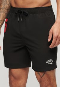 Zwarte zwembroek met een elastische tailleband, trekkoord en een rode geborduurde nummer '5'. Bevat witte logo-details op het been.