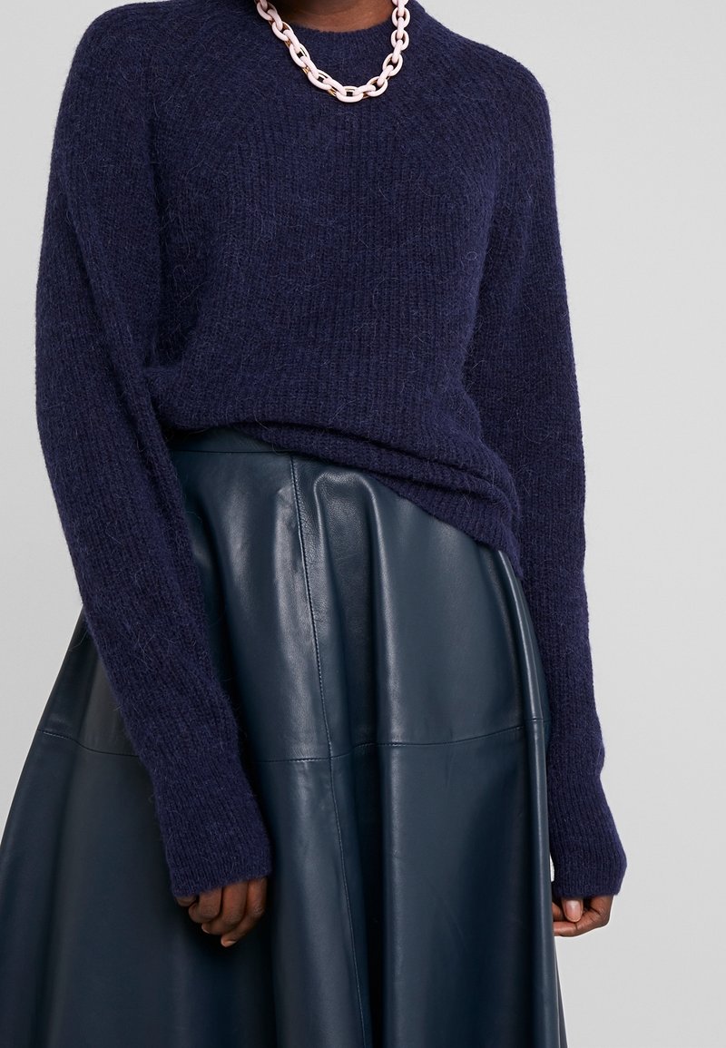 Personne portant un pull duveteux bleu marine foncé rentré dans une jupe en cuir bleu foncé, accessoirisé d'un collier à gros maillons rose clair.