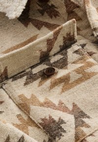 Gros plan d'une veste en laine beige avec motif géométrique marron, poche à rabat boutonnée et doublure de col douce et texturée.