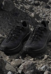 Paire de chaussures de sport noires avec lacets sur un terrain extérieur rocheux avec des pierres éparpillées et un sol irrégulier.