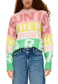 Kvinna klädd i en pastellfärgad rosa, gul och grön stickad tröja med stora blockbokstäver, tillsammans med högmidjade blåjeans.