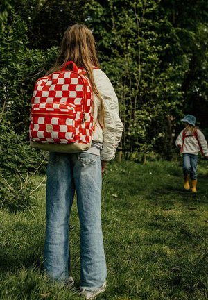 Rood-wit geblokte rugzak met ritssluiting, verstelbare schouderbanden en een beige basis; gedragen door een persoon in een lichte jas en jeans in een grasgebied.