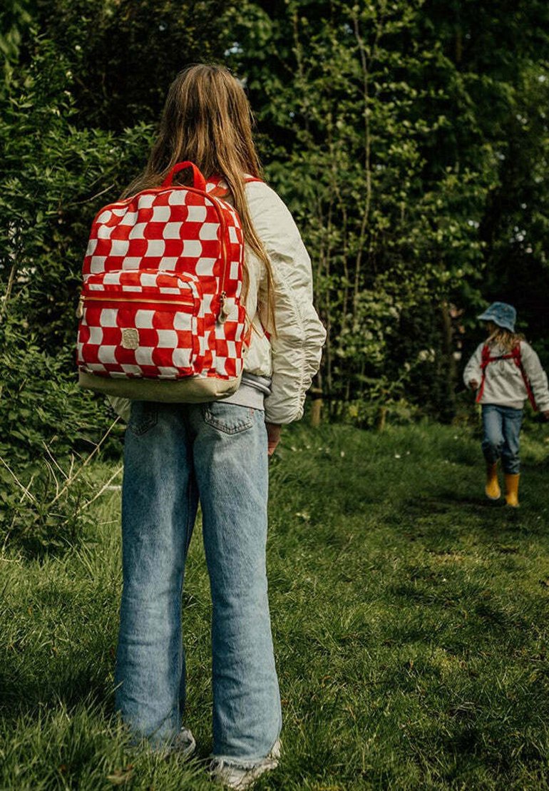 Rood-wit geblokte rugzak met ritssluiting, verstelbare schouderbanden en een beige basis; gedragen door een persoon in een lichte jas en jeans in een grasgebied.