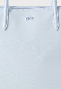 Jasnoniebieska teksturowana torba na ramię z pasującymi uchwytami i małym wyszywanym białym logo krokodyla z czerwonym detalem blisko górnego środka.