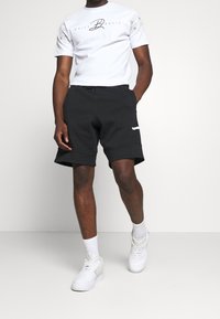 Bílý krátký rukáv tričko s tištěným logem; spárované s černými teplákovými kraťasy s bočními kapsami. Model nosí bílé tenisky.