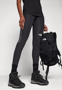 Leggings atléticas pretas com texto branco "ATHLETICS", combinados com uma mochila preta e botas de caminhada pretas resistentes. Material macio e elástico.
