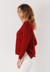Donna con capelli biondi mossi che indossa un maglione rosso a maniche lunghe e pantaloni bianchi, in piedi di profilo su uno sfondo chiaro e semplice.