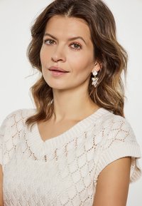 Gebreide trui in crème, met een diamanten patroon en een brede v-hals. Het model draagt bloemen-druppeloorbellen, die textuur en detail toevoegen.