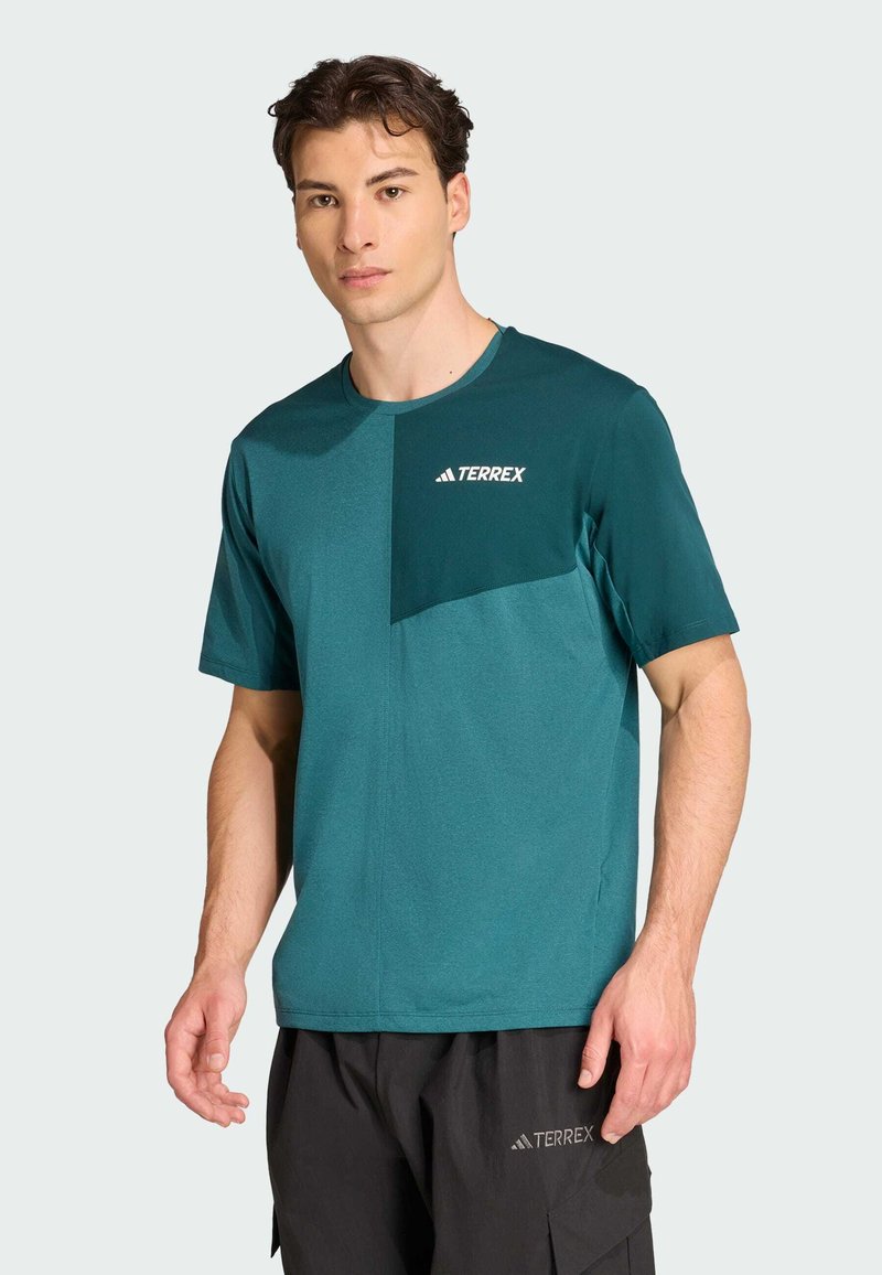 Camiseta deportiva en color azul verdoso con cuello redondo y mangas cortas; presenta un bolsillo en el pecho de contraste y el logo de Terrex en blanco en la parte superior izquierda.