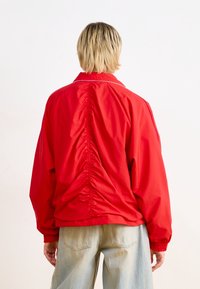 Röd jacka med avslappnad passform, featuring en samlad bakdel, elastiska ärmslut och en klassisk krage. Slät textur, minimal detaljering.