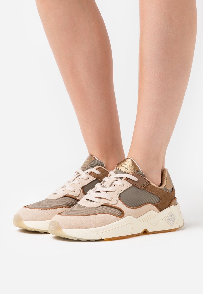Pieds portant des baskets beige et marron à lacets avec des semelles épaisses blanches sur un fond uni.