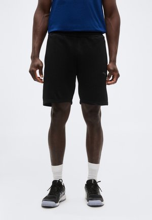 Μαύρο αθλητικό σορτς που φοράει ένα άτομο όρθιο, συνδυασμένο με λευκές κάλτσες μέχρι τον αστράγαλο, μαύρα αθλητικά παπούτσια και μπλε πουκάμισο ορατό στη μέση.