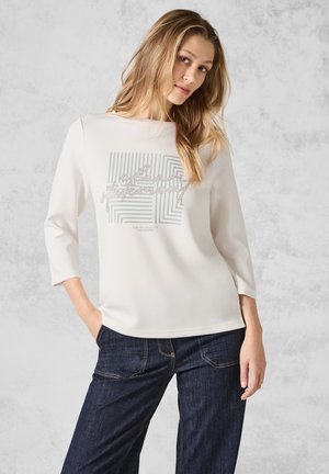 Bílý top s tříčtvrtečním rukávem se stříbrným geometrickým grafickým designem na přední části, doplněný tmavě modrými denimovými džíny.