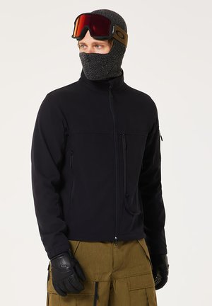 Black softshell jacket with zippered pockets, paired with a gray knit balaclava and goggles. Textured gloves complete the outdoor look.