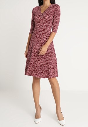 Vrouw die een knie-lange rode jurk met witte bloemenprint draagt, driekwartmouwen, V-hals en witte puntige hakken, staand tegen een effen achtergrond.