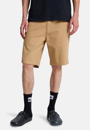 Mężczyzna ubrany w beżowe, sięgające do kolan krótkie spodenki, czarne skarpetki z białymi logo oraz czarne buty wsuwane, stojący na jednolitym tle.