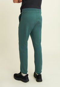 Persona que lleva pantalones de chándal color azul verdoso, zapatillas negras y una camiseta negra, de pie contra un fondo claro y liso.