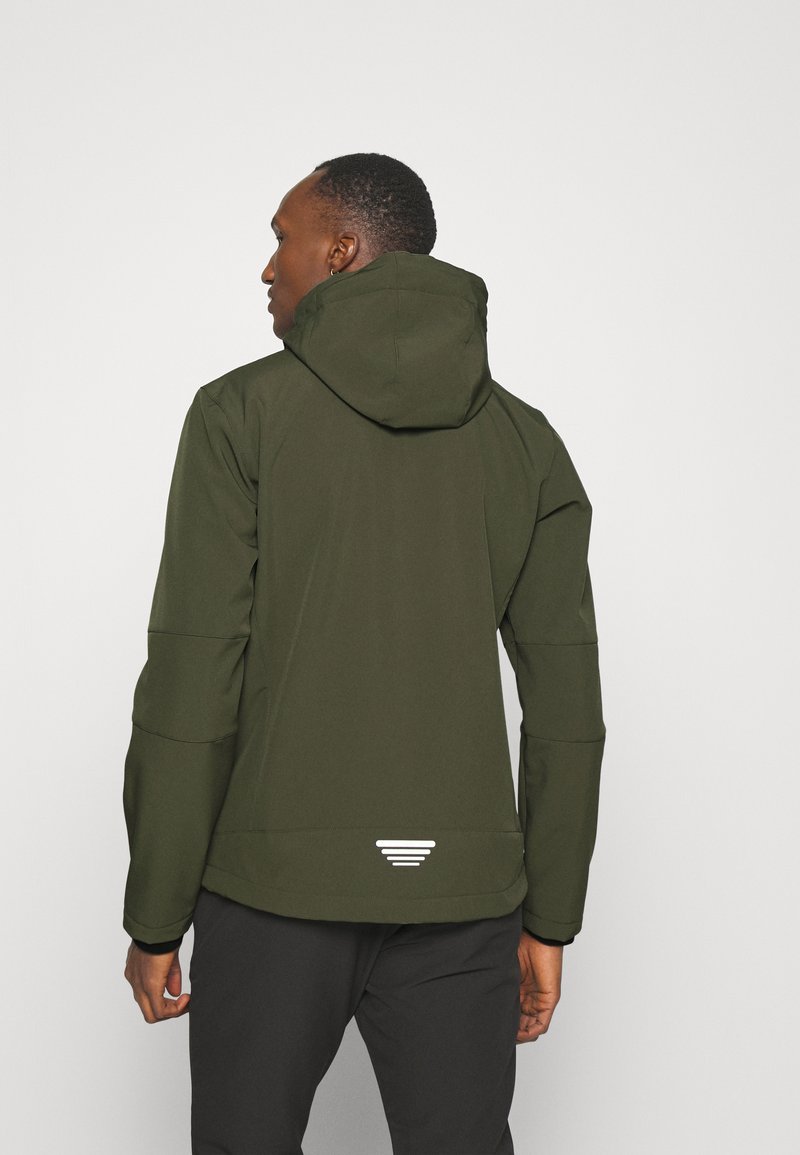 Hombre con una chaqueta con capucha verde oliva, de espaldas, con pantalones negros y un pequeño logo blanco cerca de la parte inferior de la chaqueta.