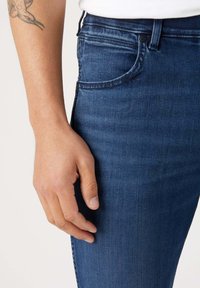 Jeans de mezclilla azul oscuro con un corte ajustado. Las características incluyen costuras sutiles, un bolsillo delantero y una mano reposando sobre el muslo. Textura suave visible.