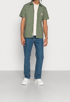 Chemise verte claire à manches courtes avec col, fermeture à boutons sur le devant, poche poitrine et deux poches avant, assortie à un jean bleu et des chaussures blanches.