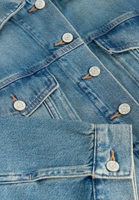 Gros plan sur une veste en denim bleu montrant des boutons en métal et des poches avec des coutures détaillées.