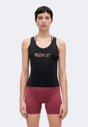 Młoda osoba z krótkimi ciemnymi włosami, ubrana w czarną koszulkę Nike na ramiączka i dopasowane bordowe spodenki sportowe, na jasnym, jednolitym tle.