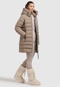 Beige Puffer-Jacke mit Kapuze, mit einem gesteppeten Design, frontalen Reißverschlusstaschen und eng gerafften Bündchen. Kombiniert mit beigen Leggings und Schneestiefeln.