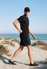 Jeune homme marchant sur des dunes de sable de la plage, portant une casquette noire, des lunettes de soleil, un polo, un short et des chaussures noires avec l'océan en arrière-plan.