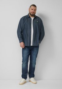 Donkerblauw denim overhemd met knopen en een borstlogo, gedragen over een wit T-shirt. Gecombineerd met een blauwe spijkerbroek en witte sneakers.