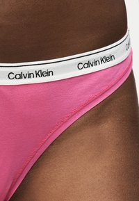 Gros plan d'une personne à la peau foncée portant des sous-vêtements roses Calvin Klein avec une ceinture blanche arborant le nom de la marque en lettres noires.