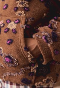 Brązowa marynarka z filcu z fioletowymi i beżowymi kwiatowymi zdobieniami. Posiada dopasowany krój oraz kieszenie, podkreślając fakturę i głębię.
