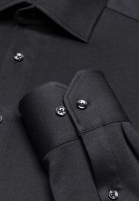 Camicia nera a maniche lunghe con colletto abbottonato, polsini lunghi con chiusura a bottone, tessuto liscio e due bottoni metallici neri.