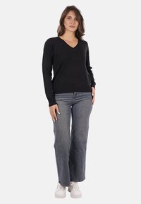 Maglione nero a V con bordi a coste, abbinato a jeans chiari a zampa d'elefante e sneaker bianche. Il modello si trova davanti a uno sfondo neutro.