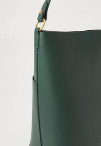 Bolsa de couro verde escura com detalhe de anel metálico dourado e alça ajustável, sobre um fundo branco.
