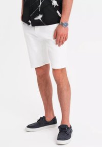 Pantalones cortos blancos con un corte a medida y bolsillos laterales, combinados con zapatos de lona azul oscuro con suelas blancas. Camisa floral en blanco y negro visible.