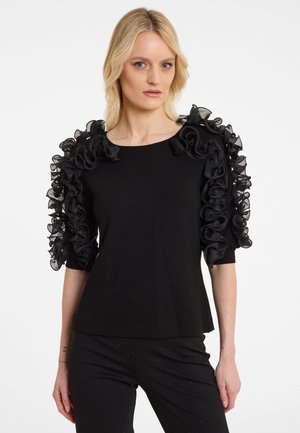 Zwarte top met fladdermouwen van gerimpeld doorschijnend stof. Eenvoudige ronde halslijn en een ontspannen pasvorm. Contrast met donkere broek met versieringen.