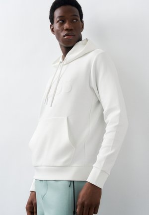 Moški, oblečen v belo jopo z žepom za roke in svetlo modre hlače, gleda levo na enobarvnem belem ozadju.